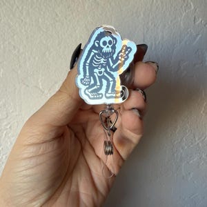 May include: A holographic badge reel featuring a skeleton-like creature giving a peace sign. The badge reel has a clear plastic retractable cord and a metal clip. The design is a light blue color and is held in a hand.