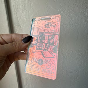 May include: A rectangular, iridescent acrylic badge featuring a graphic illustration of a flight dispatch workstation. The design includes a helicopter, computer monitors, a checklist, and the text "FLIGHT DISPATCH" and "THE FLIGHT DISPATCH".