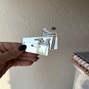May include: Silver-colored skeleton-shaped earrings displayed on a white card. The earrings feature a detailed skeleton design. The card has a rectangular shape with a hole at the top for hanging. The background is a light-colored wall.