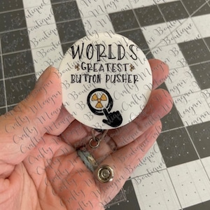 May include: A white button with a black and yellow radiation symbol and the text "World's Greatest Button Pusher" on it. The button is attached to a silver retractable badge holder.