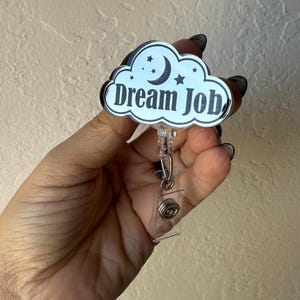 May include: A badge reel with a cloud-shaped design. The cloud is white with a black border and features a crescent moon, stars, and the words "Dream Job" in a bold, black font. The reel has a clear plastic strap and a metal clip.