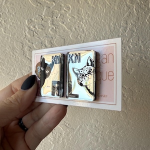 May include: Two rectangular mirrored pins featuring cat illustrations and the letters KN and RL. The left pin shows a black and white cat profile, while the right pin displays a detailed cat face. Both pins are mounted on a white display card.