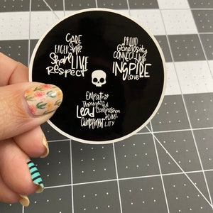 May include: A black and white round sticker with a skull in the center and words around it, including "Care", "Eager", "Smile", "Share", "Live", "Respect", "Proud", "Generosity", "Connect", "Help", "Inspire", "Love", "Empathy", "Thoughtful", "Lead", "Compassion", "Humility", and "Compliment".