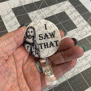 May include: A white button badge with a black and white image of Jesus Christ and the text "I SAW THAT". The badge is attached to a clear plastic retractable badge holder.
