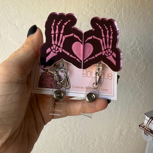 May include: A pair of pink and black badge reels featuring a skeleton hand heart design. The badge reels have a clear plastic retractable cord and a metal clip. The card behind the badge reels has the text "Boutique Custom Radiology Gear".