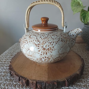 May include: Cream ceramic teapot with a brown floral design and a matching lid. The teapot has a woven handle and is displayed on a wooden slice. The teapot is a decorative item.