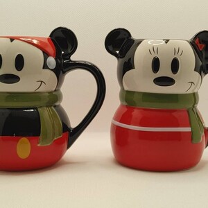 May include: Two ceramic mugs, one depicting Mickey Mouse in a Santa hat and green scarf, the other Minnie Mouse with a red bow and scarf. Both mugs have black handles and are red, black, and white.