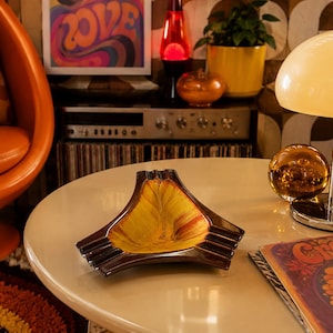 May include: A triangular ceramic ashtray featuring a glossy dark brown outer rim and a vibrant yellow and orange marbled centre. The piece has three distinct notched rests for cigarettes, showcasing a smooth, reflective finish on its surface.