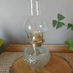 May include: A clear glass oil lamp featuring a textured diamond pattern on the round base, a brass burner assembly with a small adjustment knob, and a tall, transparent glass chimney globe that tapers toward the top for a classic lighting design.
