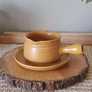 May include: A mustard yellow ceramic sauce boat with a single handle and a matching saucer. The sauce boat features a series of horizontal ridges around its base and a small pouring spout. The set has a smooth, matte finish throughout.