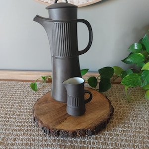 May include: A dark brown ceramic coffee pot with a lid and a small matching creamer. The coffee pot has vertical ribbed detailing and a curved handle. The creamer has a small handle and a white interior. Both are displayed on a wooden slice.