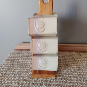 Peut inclure: Un porte-savon en céramique empilé avec une poignée en bois. Le porte-savon est composé de trois blocs rectangulaires, chacun avec un motif de soleil et un dégradé de couleurs rose, bleu et blanc. La poignée en bois a un trou circulaire.