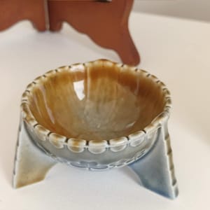 Peut inclure: Un petit bol en céramique brun et bleu avec trois pieds. Le bol a un bord festonné et un intérieur blanc.