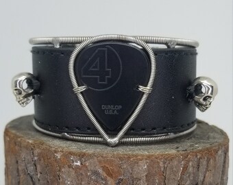 Könnte beinhalten: Schwarzes Lederarmband mit einem silbernen Gitarrenplektrum, Totenkopf-Anhängern und dem Text "Dunlop U.S.A." auf dem Plektrum.