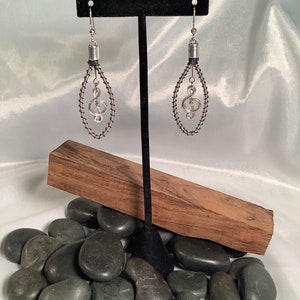 May include: Silver treble clef earrings with black wire wrapped around the loop. The earrings are hanging from a black stand on a bed of black river rocks.