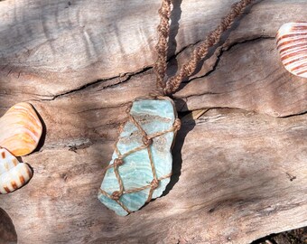 Pode incluir: Um pingente de cristal bruto azul claro e branco preso em uma rede de cordão de macramê marrom. O pingente pende de um colar de cordão de macramê marrom correspondente, apresentando um padrão de nós em espiral que se estende para cima.