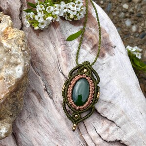 May include: A handmade macrame necklace featuring a dark green oval stone pendant. The pendant is framed with olive green and tan macrame, accented with small gold beads. The necklace is displayed on a piece of driftwood, with white flowers.