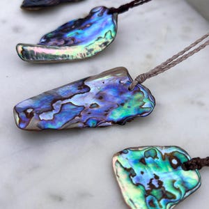 May include: Four iridescent abalone shell pendants with natural blue, green, and purple hues. Each shell is unique in shape and size, strung on a brown or tan cord. The pendants are displayed on a white marble surface.
