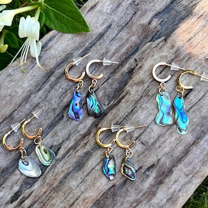 Puede incluir: Varios pares de pendientes de aro con colgantes de concha de abulón iridiscente. Los pendientes tienen aros y postes de color dorado. Las conchas de abulón muestran una gama de colores, incluyendo azul, verde y púrpura. Los pendientes se exhiben sobre una superficie de madera envejecida.