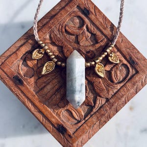 May include: A necklace featuring a light-colored crystal pendant with a hexagonal shape. The pendant is suspended from a braided cord and adorned with gold-colored leaf and bead accents. The necklace rests on a carved wooden surface.