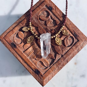 May include: A clear quartz crystal pendant on a brown cord necklace with gold accents. The necklace is displayed on a wooden box with intricate carvings.