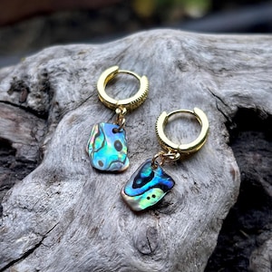 May include: Gold hoop earrings with iridescent abalone shell pendants. The shell pieces have blue, green, and purple colours. The earrings are shown on a piece of weathered wood.