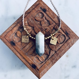 May include: A white crystal pendant necklace with gold accents and beads. The necklace is displayed on a wooden box.