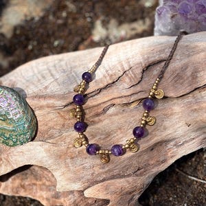 May include: A handcrafted necklace featuring deep purple amethyst beads and small gold-colored charms. The necklace is strung on a brown cord, with a natural wood background and a hint of sunlight.