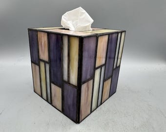 May include: A square tissue box with a stained glass design. The box features a geometric pattern of rectangular shapes in shades of purple, pink, yellow, and white. A white tissue is visible at the top.