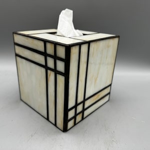 May include: A white and brown stained glass tissue box cover with black metal accents. The box is square and has a geometric design.