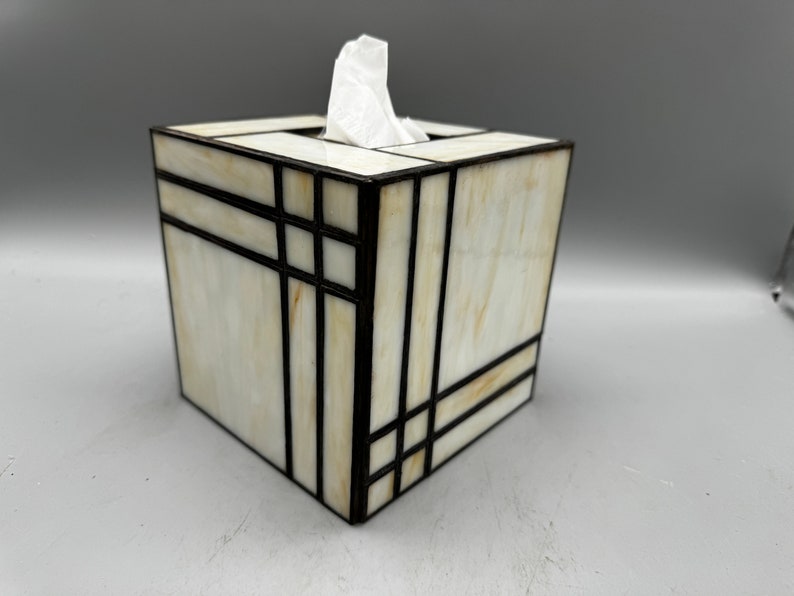 May include: A white and brown stained glass tissue box cover with black metal accents. The box is square and has a geometric design.