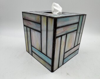 Könnte beinhalten: Ein quadratischer Taschentuchbox-Bezug mit einem Buntglasdesign. Der Bezug hat ein geometrisches Muster in Pastellfarben, darunter Blau, Rosa und Gelb, mit schwarzen Linien, die die Glasteile trennen. Oben ist ein weißes Taschentuch zu sehen.