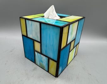 Op de afbeelding: Een tissuebox van glas in lood met een geometrisch ontwerp. De doos heeft een mix van blauwe, gele en witte glazen panelen, waardoor een kleurrijke en artistieke uitstraling ontstaat. De tissuebox is vierkant en heeft een zwarte rand.