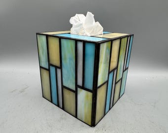 Op de afbeelding: Een vierkante tissuebox met een glas-in-lood ontwerp. De doos heeft een geometrisch patroon van gele, blauwe en witte glazen panelen, omlijnd met zwarte lijnen. Witte tissues zijn zichtbaar aan de bovenkant.