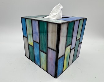 Op de afbeelding: Een tissuebox van glas in lood met een geometrisch ontwerp. De doos heeft rechthoekige panelen in blauw, groen, geel en paars, omlijst door zwart metaal. Witte tissues zijn zichtbaar aan de bovenkant.