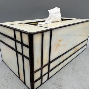 May include: Rectangular tissue box with a stained glass design. The box has a cream-coloured background with brown geometric lines. White tissues are visible at the top.