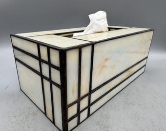 May include: Rectangular tissue box with a stained glass design. The box features a cream-colored background with brown geometric lines. White tissues are visible at the top.