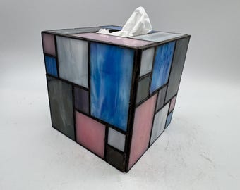 Op de afbeelding: Een vierkante tissueboxhouder gemaakt van glas-in-loodpanelen in tinten blauw, roze, wit en grijs. De glasstukken zijn verbonden door donkere metalen soldeerlijnen, wat een geometrisch mozaïekpatroon vormt met een opening aan de bovenzijde.