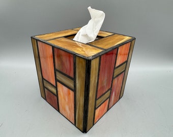May include: A square tissue box cover made of stained glass with a geometric pattern of brown, orange, and pink glass pieces. The box is empty and a white tissue is sticking out of the opening.