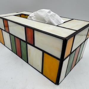 Puede incluir: Una caja para pañuelos rectangular construida con paneles de vidrio tintado unidos por líneas de soldadura de metal oscuro. El diseño presenta un patrón geométrico de rectángulos de vidrio blanco opaco, amarillo, naranja y verde translúcido.
