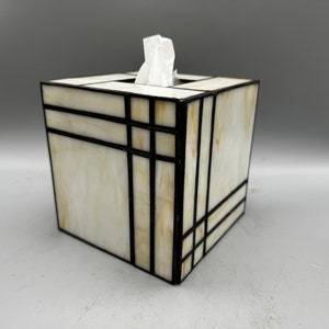 May include: A white tissue box cover with a geometric design in black and gold. The cover is made of stained glass and has a square shape.