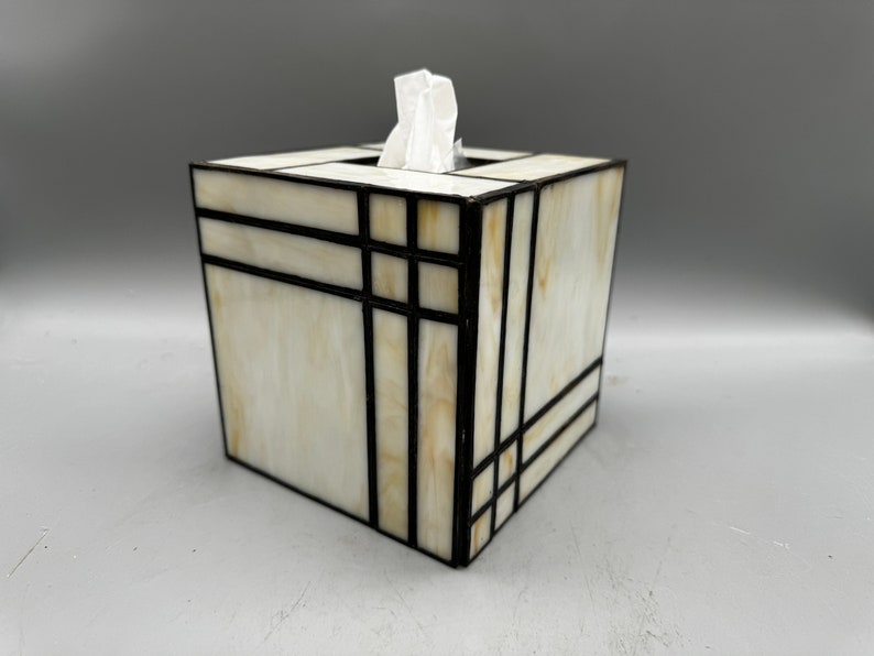 May include: A white tissue box cover with a geometric design in black and gold. The cover is made of stained glass and has a square shape.