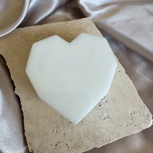 May include: A white, heart-shaped object with a geometric design rests on a beige, textured stone. The stone sits atop a shimmering, satin-like fabric in a neutral tone. The heart shape is smooth and opaque.