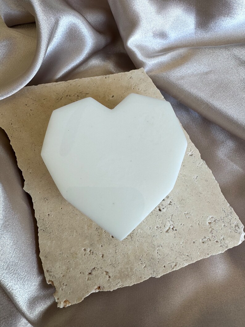May include: A white, heart-shaped object with a geometric design rests on a beige, textured stone. The stone sits atop a shimmering, satin-like fabric in a neutral tone. The heart shape is smooth and opaque.