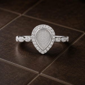 May include: A silver-toned ring featuring a pear-shaped setting with a textured, matte center. The pear-shaped halo and the band are adorned with small, clear, round-cut stones, creating a sparkling effect along the entire length of the ring.