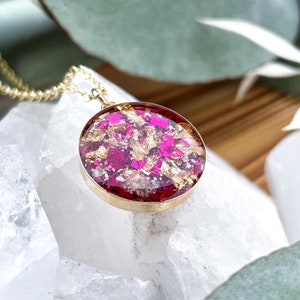 May include: A gold necklace featuring a circular pendant. The pendant is filled with pink and gold flakes set in clear resin. The necklace is displayed on a white, crystalline surface, with green foliage in the background.