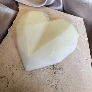 May include: A geometric heart-shaped object in a pale, creamy color. The heart has a faceted design and sits on a textured, light-colored stone. A silky, silver-gray fabric is in the background.