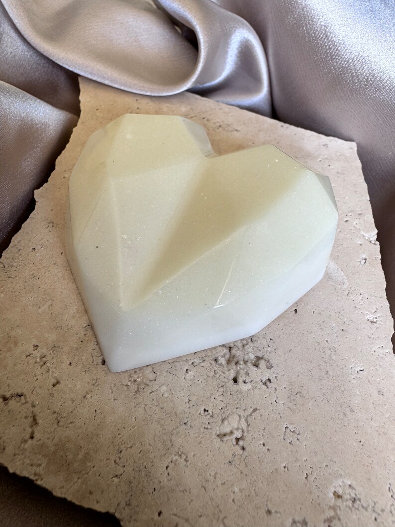 May include: A geometric heart-shaped object in a pale, creamy color. The heart has a faceted design and sits on a textured, light-colored stone. A silky, silver-gray fabric is in the background.