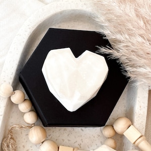May include: A white, geometric heart-shaped object rests on a black hexagonal box. The heart has a textured surface. The box sits in a speckled white tray, with wooden beads and dried ornamental grass in the background.