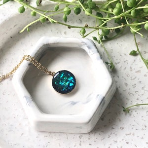 May include: A gold-toned necklace with a round pendant featuring iridescent blue and green fragments. The pendant is set in a gold frame. The necklace rests in a white and gray marble-patterned hexagonal dish.